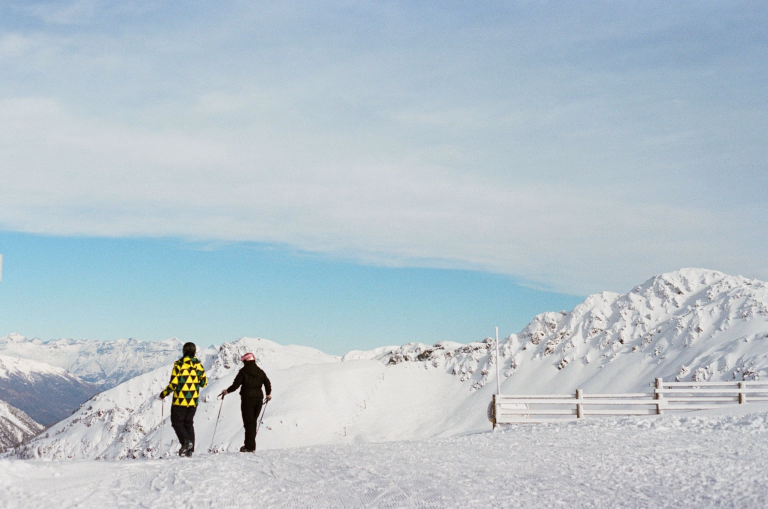 Deux personnes sur une piste de ski avec leur ski au pied, de dos, regarde les montagnes blanches