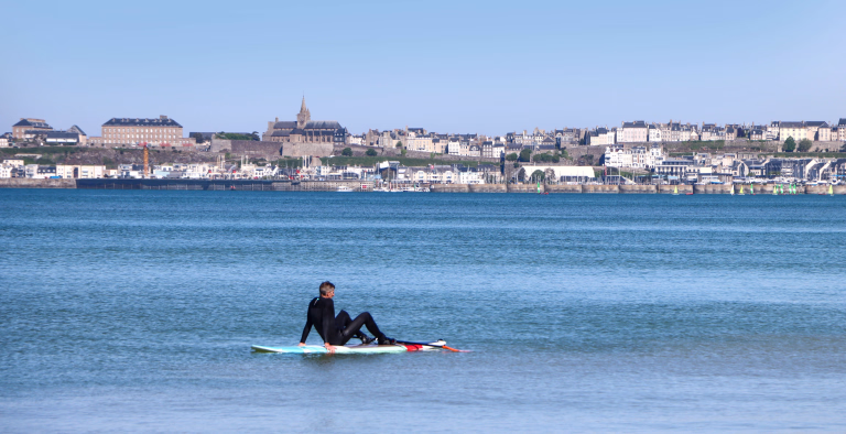 Personne assise sur une planche de surf au milieu de l'océan avec une ville côtière en arrière plan